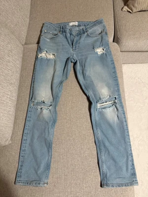Ljusblå slitna jeans med hål - Säljer ett par ljusblå jeans med slitningar och hål på både lår och knän. Jeansen har fem fickor, raka ben och klassisk passform. Perfekta för en avslappnad och trendig look. Materialet är denim i bomull.