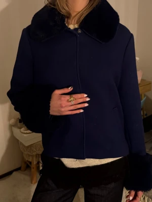 Mörkblå kort jacka med fuskpäls från Meotine - Snygg mörkblå jacka med krage och muddar i fuskpäls. Kortare modell som stängs med knappar framtill. Perfekt för dig som vill ha en stilren och trendig look till hösten eller vintern.