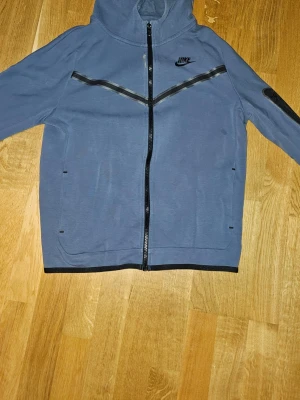Blå zip hoodie Nike tech - Snygg blå nike tech från Nike med hel dragkedja framtill och svarta detaljer. Hoodien är i strl 158-170 cm. Den har blivit blekt på vissa ställen, men det är inget man märker tydligt.