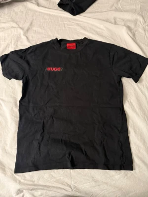 Svart t-shirt från Hugo med logga - Snygg svart t-shirt från Hugo med röd logga tryckt på bröstet. Klassisk passform med rund hals och korta ärmar. Tillverkad i mjuk bomull som känns skön mot huden. Perfekt till jeans eller shorts för en clean och enkel look.