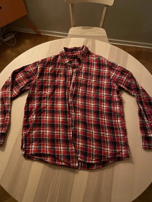 Röd och svart rutig flanellskjorta - Klassisk rutig skjorta i rött, svart och vitt med lång ärm och knappar framtill. Skjortan har en avslappnad passform och bröstficka. Perfekt för en avslappnad och cool stil. Strolek L och sitter som M-L