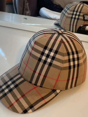 Beige rutig keps från Burberry - Säljer en klassisk beige keps från Burberry med det ikoniska rutmönstret i svart, vitt och rött. Kepsen har böjd skärm och justerbar rem bak. Perfekt accessoar för dig som vill ha en tidlös och trendig look. Storlek L 