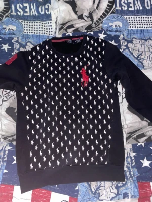 Svart tröja Polo Ralph Lauren - Svart sweatshirt från Polo Ralph Lauren med vit logotypmönster över hela framsidan och stor röd Polo-logga på bröstet. Rund halsringning och ribbade muddar. Tillverkad i bomull för skön känsla.