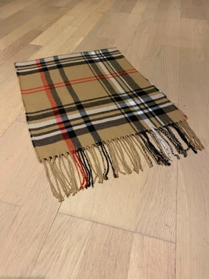 Säljer en halsduk i färgen beige.  - Säljer en Burberry halsduk i färgen Beige och röd/svarta ränderr. Den är helt ny, och oanvänd . 