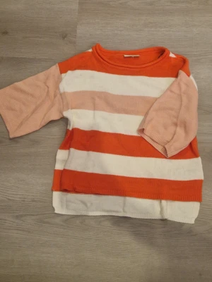 Randig stickad tröja i orange och rosa - Stickad tröja med breda horisontella ränder i orange, ljusrosa och vitt. Tröjan har rund halsringning och trekvartsärmar i ljusrosa. Har aldrig andas vilket inte är min typ på tröja. 