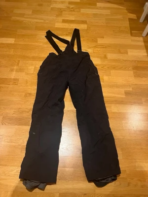 Svarta skidbyxor med hängslen - Säljer ett par svarta skidbyxor med justerbara hängslen och dragkedjor vid fickorna och vid midjan. Byxorna har en rak passform och är perfekta för skidåkning. Passar den som är runt 165cm lång. Materialet är slitstarkt och vattenavvisande, vilket gör dem grymma för vinterns alla äventyr. Är från Everest