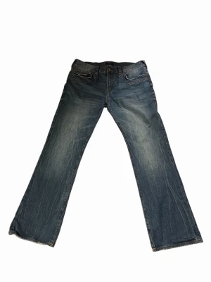 Blå bootcut jeans från True Religion - Säljer ett par blå bootcut jeans från True Religion med slitningar och ljusare partier framtill och baktill. Jeansen har klassiska fem fickor, knappgylf och dekorativa sömmar. Materialet är denim i bomull och passformen är avslappnad med utsvängda ben.