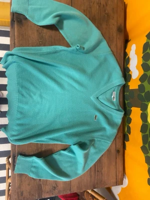 Turkos stickad tröja från Lacoste - Snygg turkos stickad tröja från Lacoste med klassisk v-ringning och den ikoniska krokodilloggan på bröstet. Tröjan har långa ärmar och ribbade muddar. Materialet är mjukt och tunt stickat, perfekt för lager på lager. Tycker att färgen blev mer blå på första bilden och att den andra bilden representerar färgen bättre