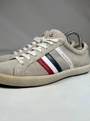 Moncler sneakers i mocka med ränder - Ja köpte dom för 6350kr helt nya. De är jätte bra skick och Dom är helt nya jätte bra skick.Snygga sneakers från Moncler i beige mocka med klassiska ränder i vitt, blått och rött på sidan. Skorna har rund tå, platt sula och vita snören. Moncler-logga på sidan och tungan ger en exklusiv känsla. Perfekta för dig som gillar stilrena och sportiga sneakers.