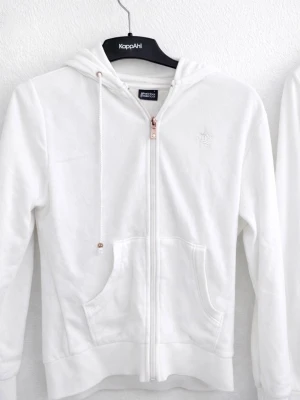 Vit hoodie från Gina Tricot - 2 st i samma storlek. Klassisk vit hoodie från Gina Tricot med dragkedja framtill, två stora fickor och justerbar huva med snören. Tillverkad i mjukt material och har en liten broderad krona på bröstet. Perfekt för en avslappnad och stilren look. 