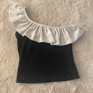 Svart oneshoulder topp med volang - Snygg svart oneshoulder topp med vit volang längs halsringningen.