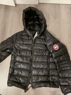  Canada Goose jacka  - Säljer en svart dunjacka från Canada Goose med huva och klassisk logotyp på ärmen. Jackan har quiltad design, dragkedja framtill och två sidofickor. Perfekt för kalla vinterdagar och riktigt varm tack vare dunfyllningen.