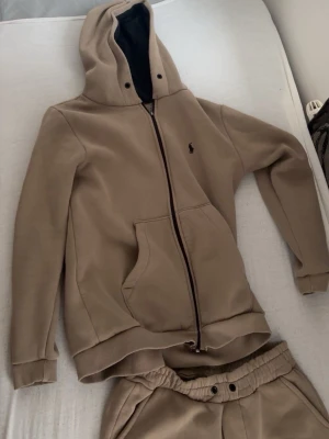 Beige loungewear-set från Polo Ralph Lauren - Säljer ett beige loungewear-set från Polo Ralph Lauren med matchande hoodie och byxor. Hoodien har dragkedja, fickor och svart logga på bröstet. Byxorna har resår i midjan och vid benslut samt Polo-logga på benet. Perfekt för chill dagar hemma.