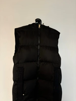 Svart dunväst med huva - Snygg svart dunväst med huva och dragkedja framtill. Västen har breda, vadderade paneler och diskreta detaljer längs sidorna. Perfekt för lager på lager och ger en clean streetwear-look.