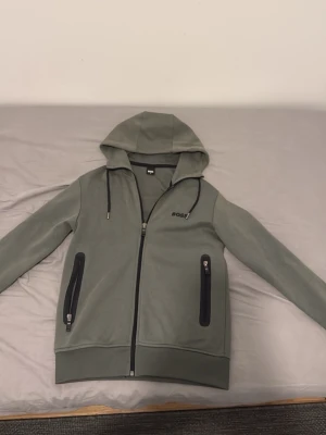 Hugo Boss hoodie - Snygg grön hoodie från BOSS med hel dragkedja framtill och två svarta dragkedjeförsedda fickor. Huva med dragsko och diskret BOSS-logga på bröstet. Tillverkad i mjukt material som känns skönt mot huden. Perfekt för en chill och stilren look. Tröjan är limiterad och är i mycket bra skick och är användbar. Tröjan är använd fåtal gånger, Inga konstigheter med tröjan dragkedjorna är inte svåra att dra ner eller up. Kom dm för frågor. passar också Xs
