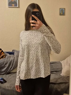 Vit prickig långärmad topp - Vit långärmad topp med svarta prickar i ett lekfullt mönster. Toppen har en rak passform och är tillverkad i ett mjukt och bekvämt material, perfekt för en avslappnad stil. Rund halsringning och diskreta slitsar i sidorna för extra rörelsefrihet. Skön tröja att ha på sig till vardags eller sova i tillochmed! 