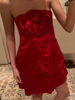 Röd capital klänning - Storlek S/M. Säljer en röd axelbandslös kort klänning med volangdetaljer och en stor rosformad dekoration framtill. Klänningen har en figurnära passform och är perfekt för capitol tema fest. Materialet känns lent och har en lätt glans. Köpt på Sellpy i mycket bra skick från Nardini Collection. Prislapp på. 