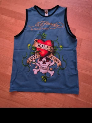 Blå Ed Hardy linnetopp med tryck - Snygg blå linnetopp från Ed Hardy med ikoniskt dödskalle- och hjärttryck samt texten 'Love Kills Slow' på framsidan. Svarta kantband runt ärmhål och halsringning. Perfekt för dig som gillar statement-plagg och streetwear vibes.