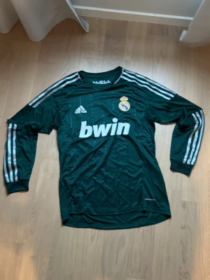 Real Madrid långärmad adidas tröja - Säljer en mörkgrön långärmad Real Madrid-tröja från adidas med vita ränder på ärmarna och klubbmärke på bröstet. Tröjan har rund hals och är tillverkad i polyester, perfekt för fotboll eller träning. Bwin-logga på framsidan.
