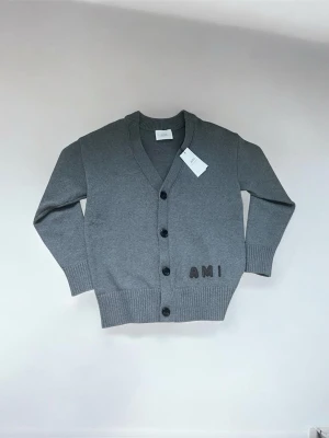 Grå cardigan från AMI Paris - Oanvänd stilren grå cardigan från AMI Paris med ribbade muddar. Perfekt för lager-på-lager och enkel att matcha. Säljer pga oanvänd födelsedagspresent. Skriv för frågor/funderingar!