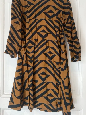 Brun och svart mönstrad klänning H&M - Cool klänning från H&M i brun och svart med grafiskt mönster. Klänningen har lång ärm, rund halsringning och är i en mjuk bomullsblandning. Passformen är normal och längden går till knäna. Perfekt för dig som gillar statement-plagg!