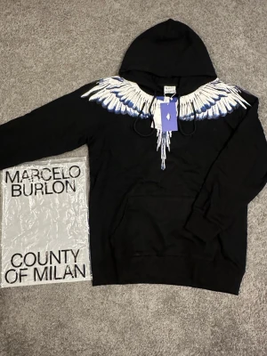Marcelo Burlon svart hoodie med vingar - HELT NY!!                                                               Svart hoodie från Marcelo Burlon County of Milan med ikoniskt vingtryck i vitt och blått över axlarna. Klassisk huva med dragsko och känguruficka framtill. Tillverkad i mjuk bomull för skön känsla. Perfekt för dig som vill sticka ut med streetwear-vibe.
