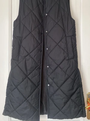 Svart lång dunväst från H&M - Stilren svart lång dunväst från H&M med quiltat mönster och tryckknappar framtill. Västen har en rak passform, rund halsringning och stora fickor på sidorna. Perfekt för lager på lager och ger en cool look till din outfit. Kan knäppas upp på sidorna nertill (se sista bilden). Storlek mer som en S
