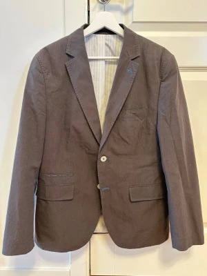 Brun kavaj från s.oliver - Stilren brun kavaj med två knappar framtill och klassisk krage. Kavajen har två fickor med lock och en bröstficka. Passar perfekt till både skjorta och t-shirt för en snygg look.