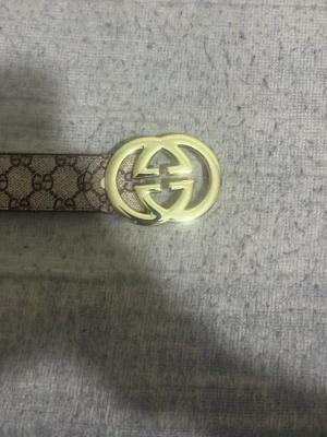 Gucci GG bälte med guldspänne - Snyggt Gucci-bälte med klassiskt GG-mönster i beige och brunt. Bältet har ett stort, glänsande guldspänne med GG-logga och är gjort i skinn. Perfekt accessoar för att lyfta din outfit.