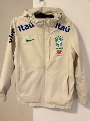 Nike Brasil vindjacka med huva - Stilren beige vindjacka från Nike med Brasilien CBF-logga, grönt swoosh-märke och sponsordetaljer i blått och rött. Jackan har hel dragkedja, huva med dragsko och två sidofickor. Perfekt för dig som gillar sportig streetwear och fotbollsinspirerad stil.