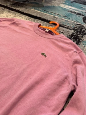 Lacoste stickad tröja - Rosa stickad tröja från Lacoste med klassisk rund halsringning och den ikoniska gröna krokodilloggan broderad på bröstet. Tröjan har långa ärmar och ribbade muddar vid ärmslut och nederkant. Perfekt för en stilren och avslappnad look.