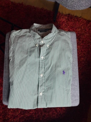 Randig skjorta från Polo Ralph Lauren - Snygg grön- och vitrandig skjorta från Polo Ralph Lauren i slim fit-modell. Skjortan har klassisk krage, knappar framtill och en broderad lila logga på bröstet. Perfekt för dig som gillar stilrena och tidlösa plagg.