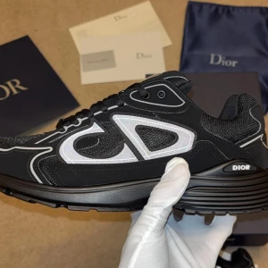 Svarta Dior sneakers med vit logga - Snygga svarta Dior sneakers med vit CD-logga på sidan. Skorna har en chunky sula, meshdetaljer och svarta skosnören. Stilen är sportig och modern med kontrasterande vita detaljer och reflekterande inslag. Perfekta för dig som gillar exklusiva och trendiga sneakers.