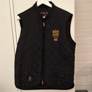 Svart quiltad väst från Morris - Stilren svart quiltad väst från Morris med broderad guldfärgad logga och brittisk flagga på bröstet. Västen har dragkedja framtill, ståkrage och diskreta fickor. Perfekt för lager-på-lager och snygg till många outfits.