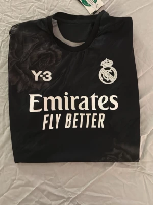 Real Madrid x Y-3 svart matchtröja - Snygg svart matchtröja från Real Madrid x Y-3 med vita tryck, klubbmärke och Emirates Fly Better-logga på bröstet. Unik rosdetalj i grått på axeln och nedre delen. Tillverkad i lätt och funktionellt material, perfekt för träning eller match.