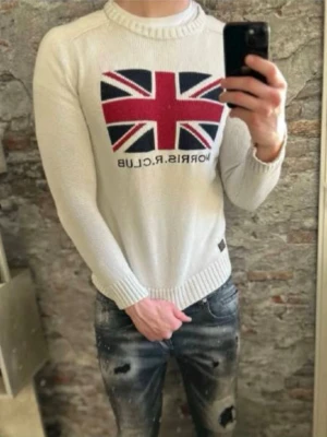 Vit stickad tröja Morris R. Club - Vit stickad tröja från Morris R. Club med Union Jack-flagga i rött, blått och vitt på bröstet. Tröjan har rund halsringning, ribbstickade muddar och är tillverkad i ett mjukt material. Perfekt för en stilren och trendig look.
