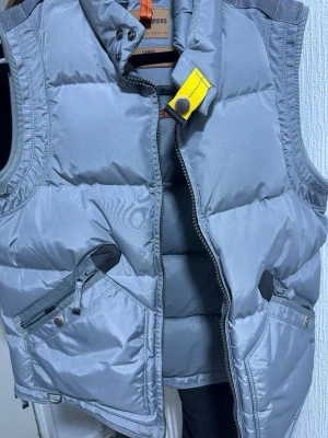 Grå dunväst från Parajumpers - Snygg grå dunväst från Parajumpers med gul detalj vid kragen och två dragkedjeförsedda fickor framtill. Västen har hög krage, quiltad design och är perfekt för lager-på-lager-stil. Klassisk look med sportig touch. Helt nytt skick, endast använd 2 gånger!