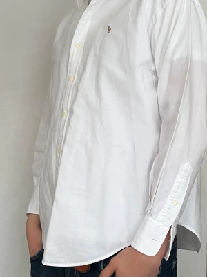 Vit skjorta från Ralph Lauren - Klassisk vit skjorta från Ralph Lauren med broderad logga på bröstet. Skjortan har lång ärm, knappar framtill och en tidlös passform. Perfekt till jeans  för en clean och stilren look. Nypris runt 1800.  Strl 12 lite för liten för mig i armarna och längd som är 188 och väger 80💵🍾👑