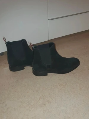 Svarta boots i mocka från Gant - Säljer ett par klassiska svarta Chelsea boots i mocka med elastiska sidopaneler och dragflikar bak. Bootsens stilrena design gör dem enkla att matcha och de har låg klack och rundad tå. Perfekta för dig som gillar tidlös och enkel stil.