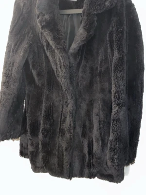 Svart fuskpälsjacka med krage - Snygg svart fuskpälsjacka med bred krage och mjukt, fluffigt material. Jackan har en rak passform och stängs framtill. Perfekt för dig som vill ha en trendig och varm look under kalla dagar.