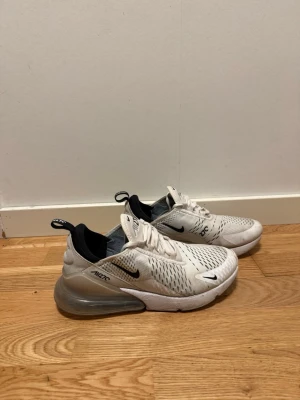 Nike Air Max 270 vita sneakers - Nike Air Max 270 sneakers i vitt med svarta detaljer och stor synlig Air-enhet i hälen. Ovandelen är i mesh för bra ventilation och har en modern, sportig look. Swoosh-logga på sidan och dragflik bak för enkel påtagning. Perfekta för dig som gillar streetwear.