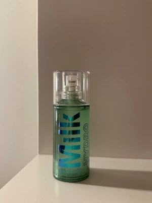 Milk Makeup Hydro Grip Primer - Säljer en Milk Makeup Hydro Grip Primer i genomskinlig plastflaska med pump och blågrön text. Primern har en gel-liknande konsistens och är perfekt för att skapa en återfuktad bas innan smink. Flaskan är cylinderformad och har ett modernt, stilrent utseende.