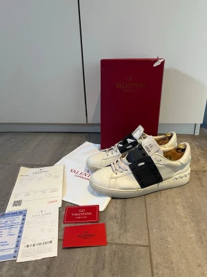 Valentino Garavani vita sneakers - Snygga vita sneakers från Valentino Garavani med svart band och nitar över sidan. Skorna har en chunky sula. Tillverkade i läder och har klassisk låg profil. Perfekta för dig som vill sticka ut med exklusiv design. Enda skadan är ett litet hål i hällen som man ser på sista bilden.