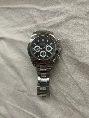 R- Daytona silverklocka - Snygg R- Daytona med svart urtavla och tre mindre kronografer. Klockan har en robust länk i silverfärgat stål och klassisk rund form. Perfekt för dig som gillar exklusiva accessoarer med sportig känsla.