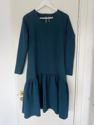 Mörk petrolblå klänning med volang - Supersnygg klänning i mörk petrolblå färg med lång ärm och rund halsringning. Modellen har en avslappnad passform och volang nedtill som ger en cool och trendig siluett. Perfekt att styla med boots eller sneakers för en modern look.