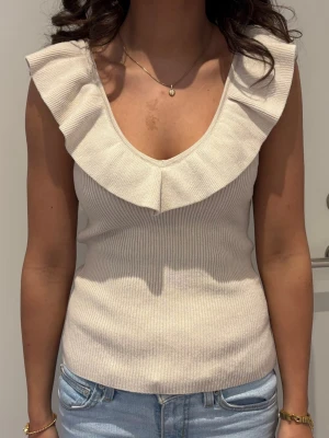 Beige ribbad ärmlös topp med volang - Supersnygg beige ribbad topp med bred volang runt både fram- och baksidan av ringningen. Toppen är ärmlös och har en djup v-ringning både fram och bak. Perfekt att styla med jeans eller kjol för en trendig look.