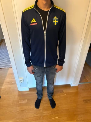 Svensk landslagsträningströja Adidas - Mörkblå träningströja från Adidas med Sveriges landslagsemblem på bröstet, gula detaljer på axlarna och vit dragkedja. Märkt med Bauhaus-logga. Perfekt för dig som vill representera Sverige på träning eller match.( T-shirt ingår )