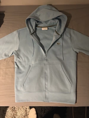 Ljusblå hoodie från Lacoste - Ljusblå hoodie från Lacoste med dragkedja framtill och klassisk krokodillogga på bröstet. Hoodien har snörning i huvan, känguruficka och är långärmad. Perfekt för en chill och stilren look.