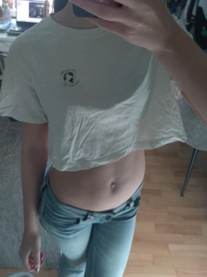 Vit cropped t-shirt med tryck - Säljer en vit cropped t-shirt med rund hals och ett litet svart tryck på vänster bröst. T-shirten har korta ärmar och är gjord i mjukt bomullstyg. Perfekt till jeans eller shorts för en avslappnad look.