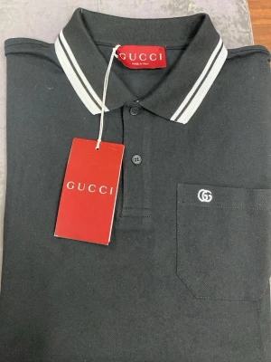 Svart pikétröja  - Klassisk svart pikétröja med vit logga broderad på bröstfickan. Tröjan har krage med vita ränder och knäppning framtill. Perfekt för en stilren och trendig look.